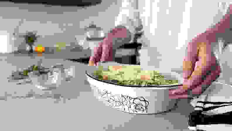 A person is trying to hold an oval-shaped Made In casserole dish with both hands. The person appears to be in a kitchen, with some ingredients, such as shredded cheese and fresh herbs, scattered in the background.