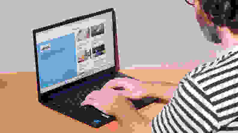 A person using an open and powered on laptop against a pastel purple background.