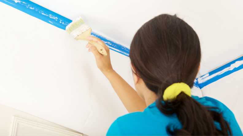 Woman using a paint brush and blue painter's tape to paint clean lines on a white ceiling.