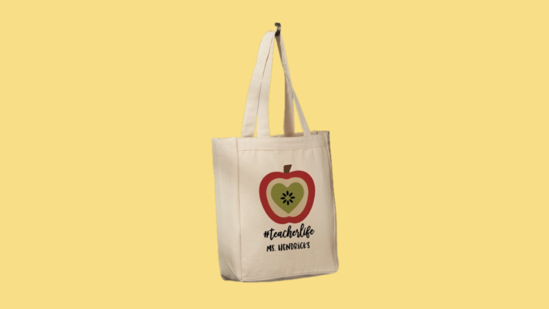 Tote bag with an apple design and text that reads "#teacherlife Ms. Hendricks" against yellow background