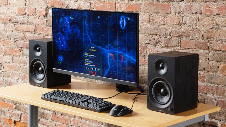 The Fluance Ai41 bookshelf speakers on either side of a computer monitor, keyboard, and mouse on a ligth wooden desk with a brick wall in the background.
