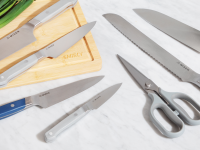 A variety of kitchen knives and a pair of sheers are spread on a marble counter and wood cutting board with scallions in the background.