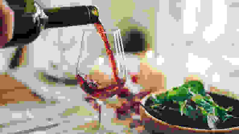 Someone pours a glass of red win beside a bowl of garden salad. A few lit candles are visible in the background, just out of focus.