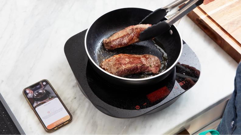 Two pieces of steak being cooked atop cookware with a smartphone displaying a recipe.