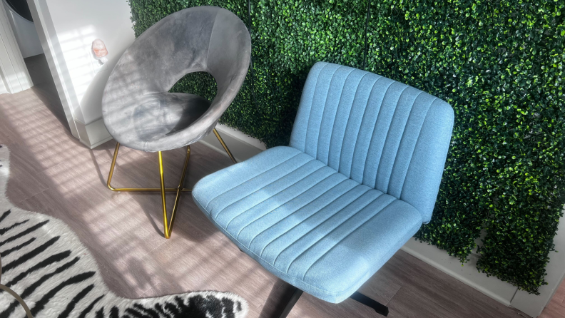 The blue Pukami Armless Desk Chair and next to gray velvet papasan desk chair sitting side by side in front of a shrubbery wall indoors on a sunny day.