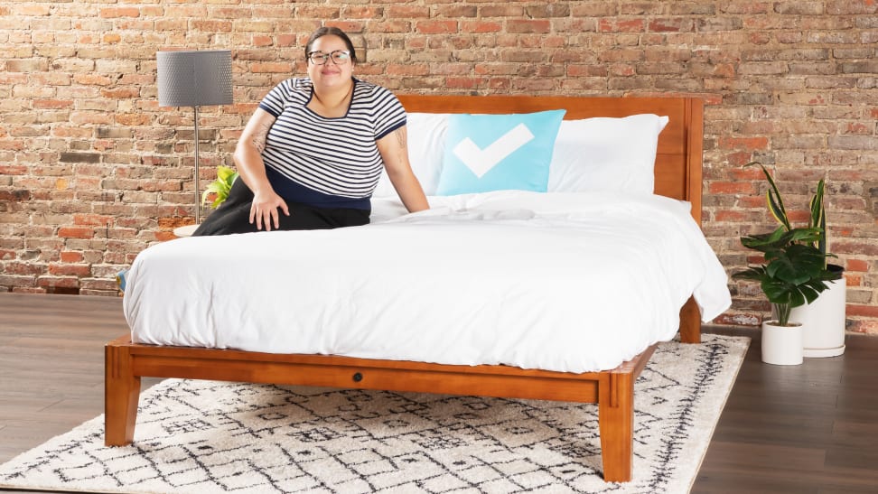 A woman sitting on a mattress atop The Bed by Thuma, a solid wood bed frame, against a brick wall.