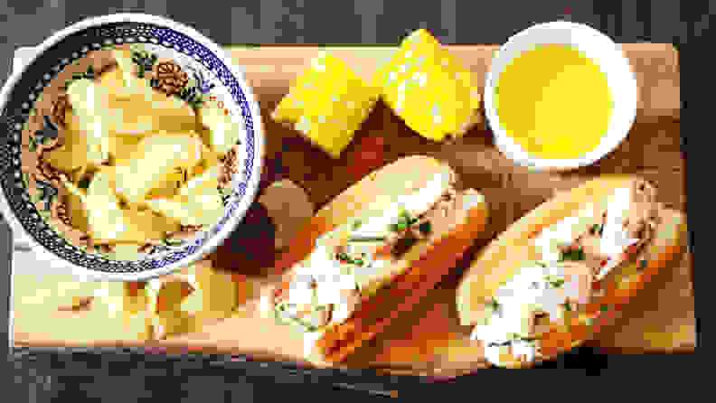 In the center of the image, there's a wooden plate and two lobster rolls on it. Next to the lobster rolls, there's a cup of melted butter, two corns, and a bowl filled with potato chips.
