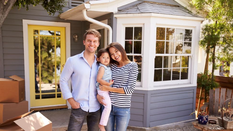 A couple with a small child stand in front of a house with moving boxes