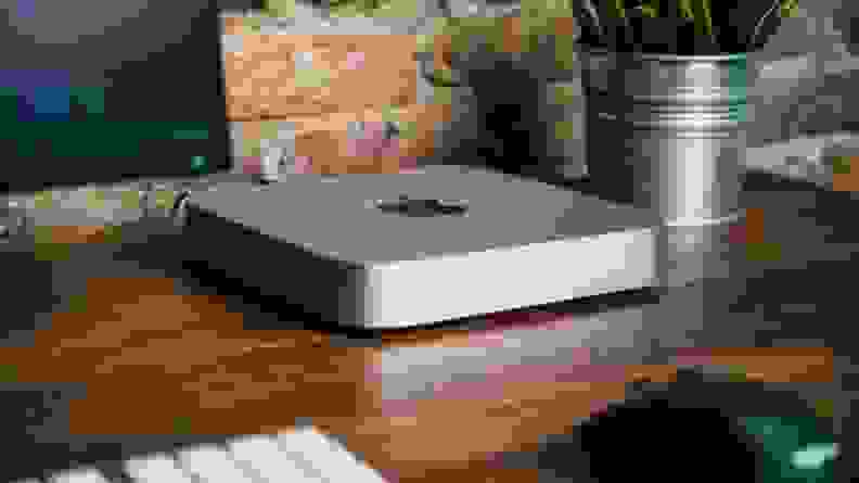 A Mac Mini sitting on a wooden desk next to a keyboard, monitor, mug, and mouse.