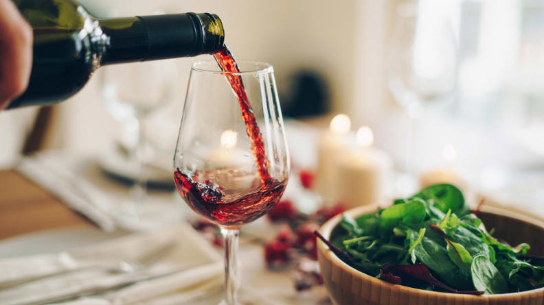Someone pours a glass of red win beside a bowl of garden salad. A few lit candles are visible in the background, just out of focus.