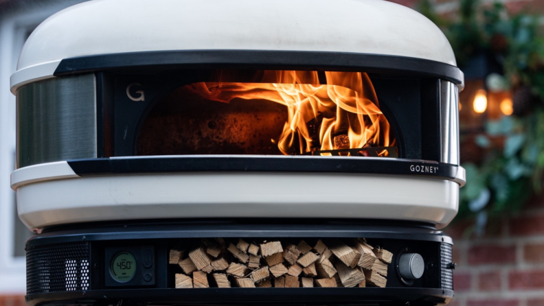 Product shot of the Gozney Dome outdoors with open flame inside and small wood stored under oven.