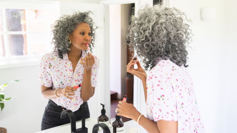 A middle-aged person with dark skin and grey hair looks into a mirror and applies a bright red liquid lipstick to her lips.