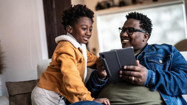 An African American father and son look at a tablet while sitting on a couch. The son wears hearing aids.