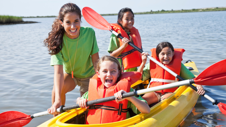 Camp counselor standing with three kids in a kayak