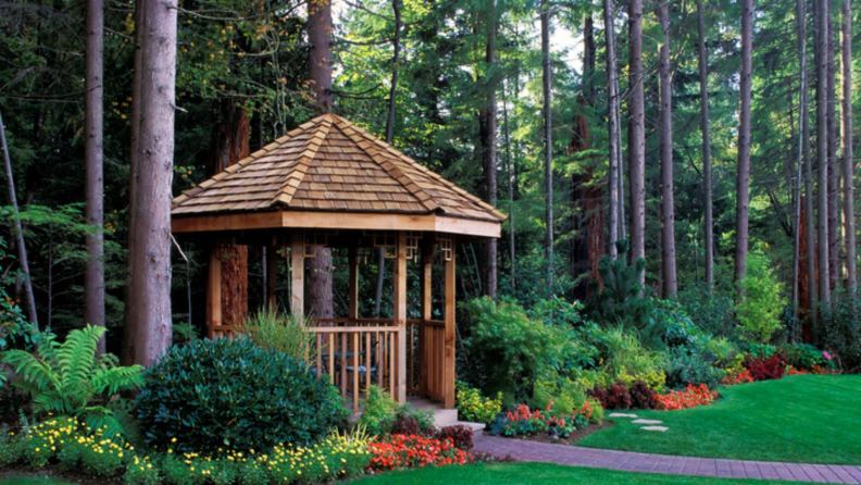 Wooden gazebo in backyard next to wooded forest area.