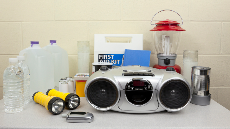 Emergency supplies on a table, including a lamp, canned food, gallon of water, flashlights, first aid kits, a flip phone, and a radio