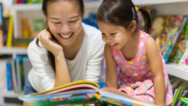 Mother and daughter reading book in library while smiling.