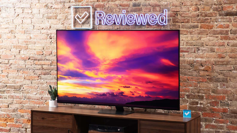 Samsung QN90D Mini-LED TV with colorful sunset on screen atop a wooden TV stand below Reviewed neon sign in front of brick wall indoors.