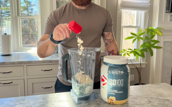 A bearded person with tattoos pours Dymatize protein powder into a shake.