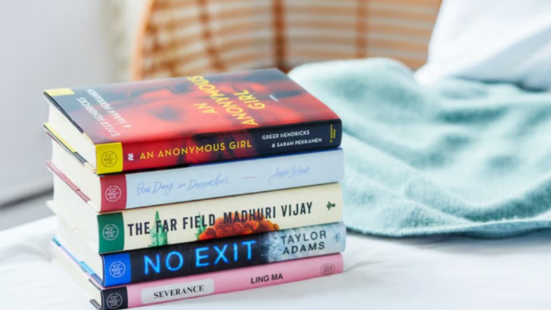 A bedroom with a stack of five books from Book of the Month resting on the bed, their spines showing the titles An Anonymous Girl, One Day in December, The Far Field, No Exit, and Severance.