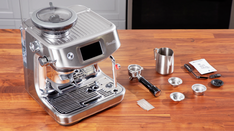 Breville Barista Touch Impress espresso machine in stainless steel finish next to stainless steel portafilter with single and dual cups filter basket, stainless steel milk frothing pitcher and a small instruction manual on top of wooden countertop surface.