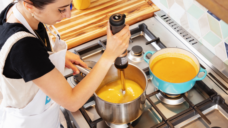 Person using a cordless immersion blender to blend butternut squash soup on a stovetop