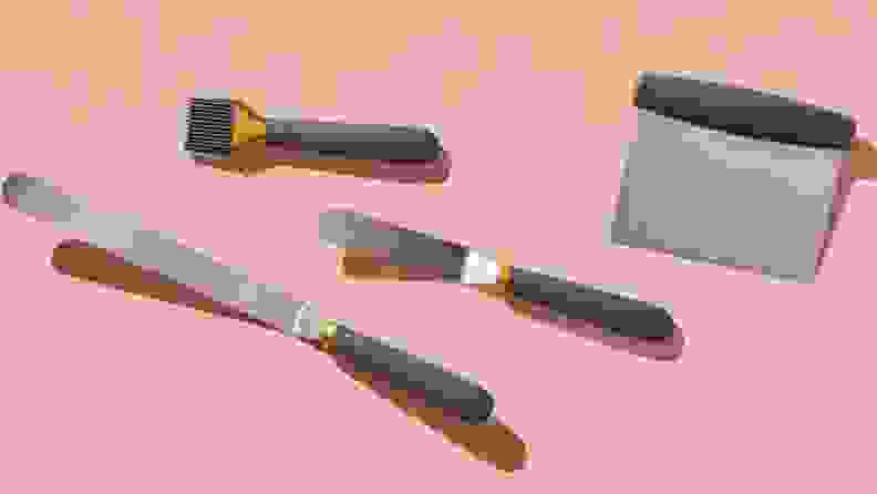 A bench scraper, small offset spatula, large offset spatula, and pastry brush, all with grey handles, laying on top of a dusty rose-colored background.