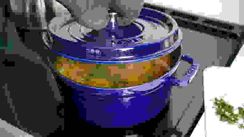 A person lifts the lid off of a blue Dutch oven to reveal chicken soup cooking inside.