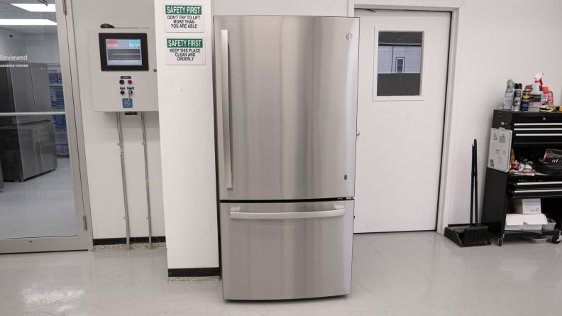 GE GDE25EYKFS Bottom-freezer Refrigerator in stainless-steel finish against white wall with safety signs and between two closed doors.