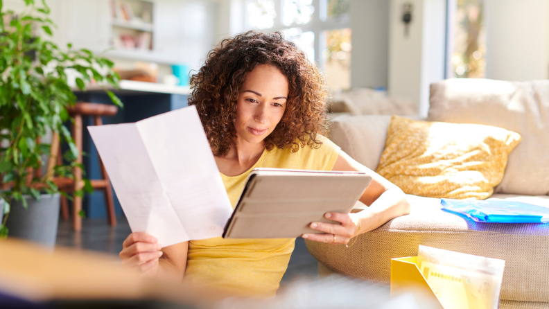 Person sitting on floor while reviewing information on paper and on laptop computer screen.