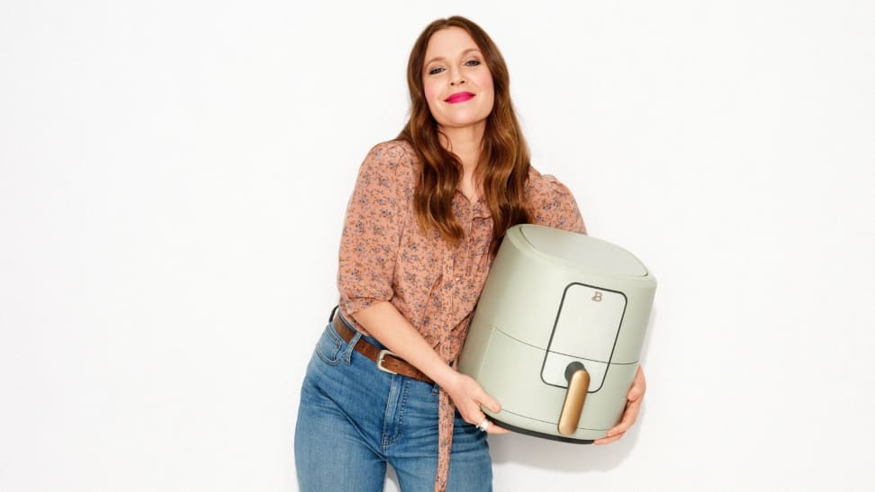 Drew Barrymore holds a sage green air fryer with a golden handle.