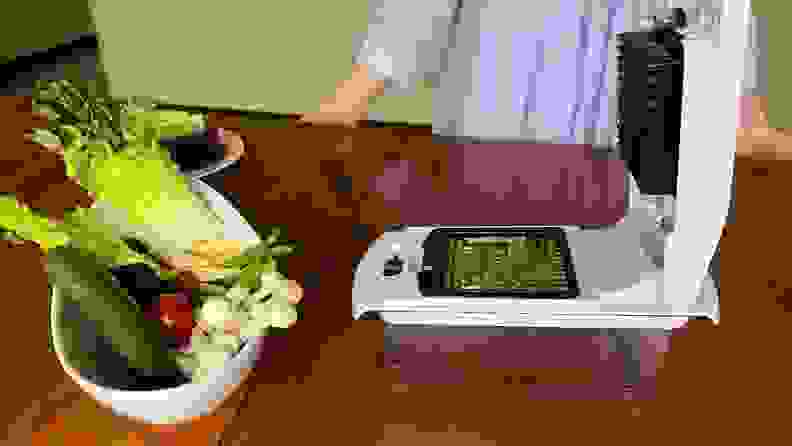Person leaning on wooden table behind several stalks of asparagus on the Fullstar Vegetable Chopper next to bowl of assorted vegetables.