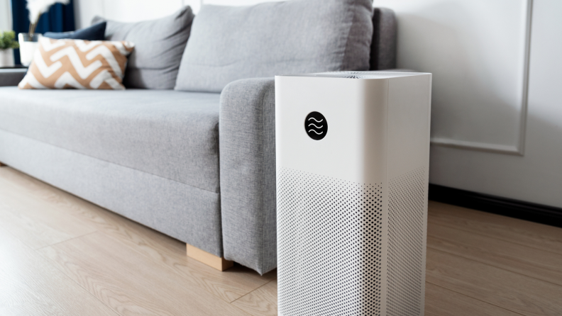 White, rectangular air purifier sitting next to gray cloth couch in living room.