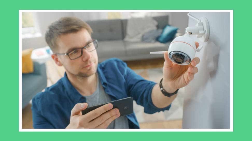 Person adjusting mounted smart security camera on wall indoors while holding smartphone.