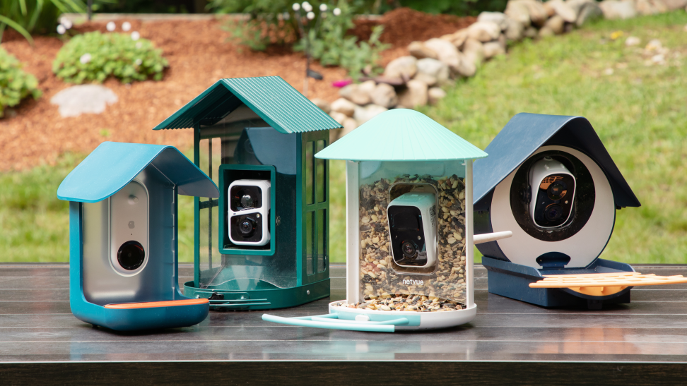 Four bird feeders in blue and green shades sit in a group on a brown table with a garden and green grass in the background