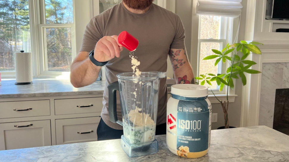 A bearded person with tattoos pours Dymatize protein powder into a shake.