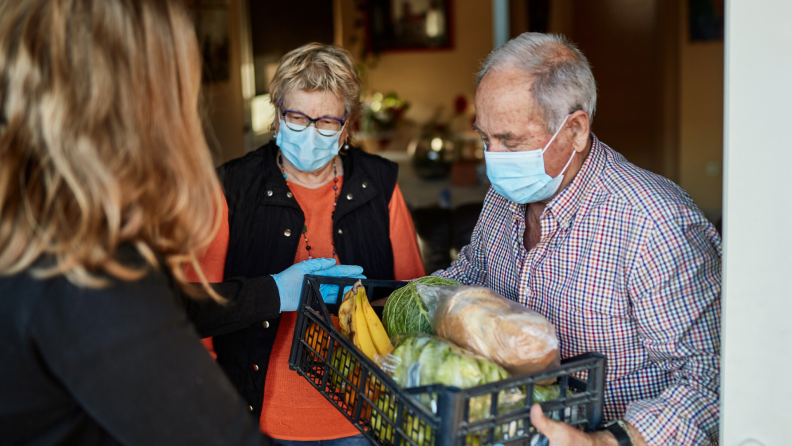 Two older adults wearing surgical masks accepting groceries in a basket from a woman.
