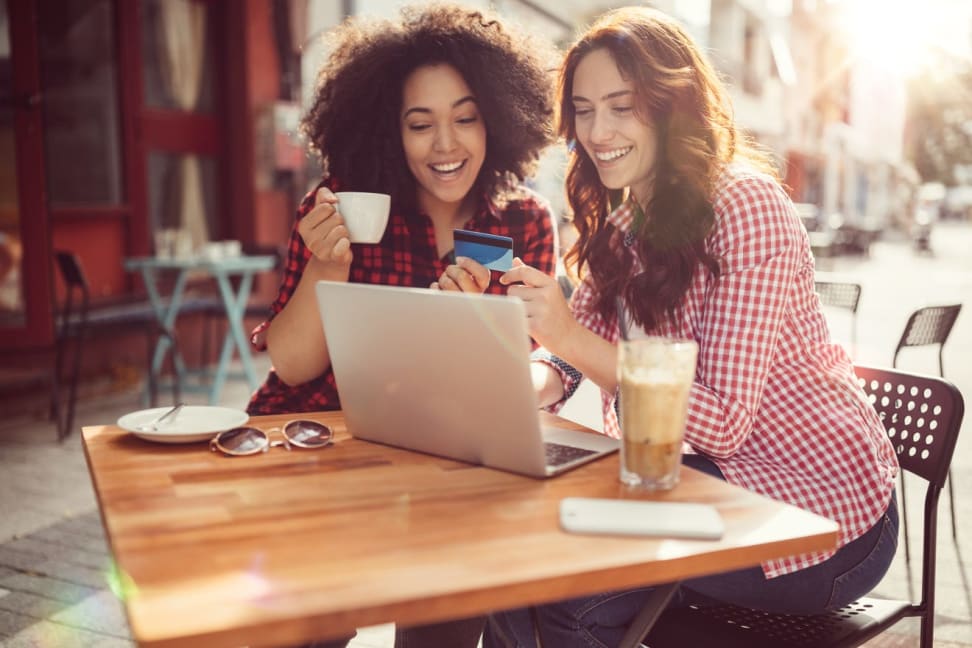 Two women sitting at table enjoying coffee while looking at laptop to shop online.