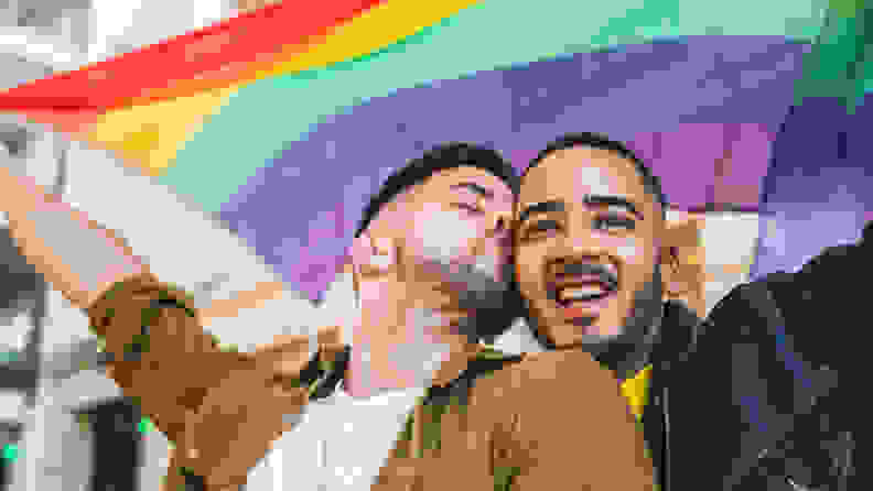 Two men holding a big rainbow flag, one kissing the other on the cheek