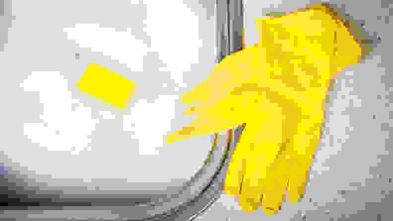 Some yellow rubber gloves sit next to a sink filled up with soapy suds. A yellow sponge is floating on top of the water, peeking out of the suds.