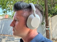 A man in a blue shirt listens to the all-white Sony WH-1000XM5. It's a bright, sunny day. The immediate setting is a residential street, but there are palm trees in hte distance.