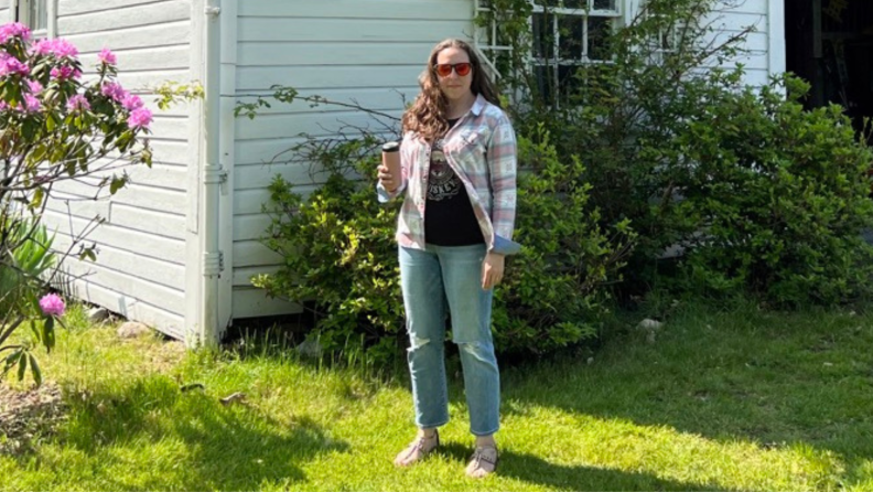 A woman stands in front of a barn holding a pink can and wearing jeans, a black t-shirt, and a plaid shirt.