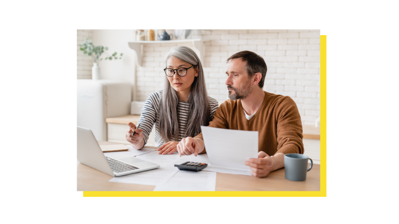 Two people sitting side by side at a table in kitchen going over paperwork and looking at computer screen.