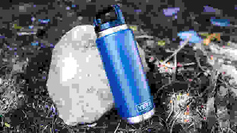 A blue Yeti Rambler water bottle leaning against a rock on the ground.