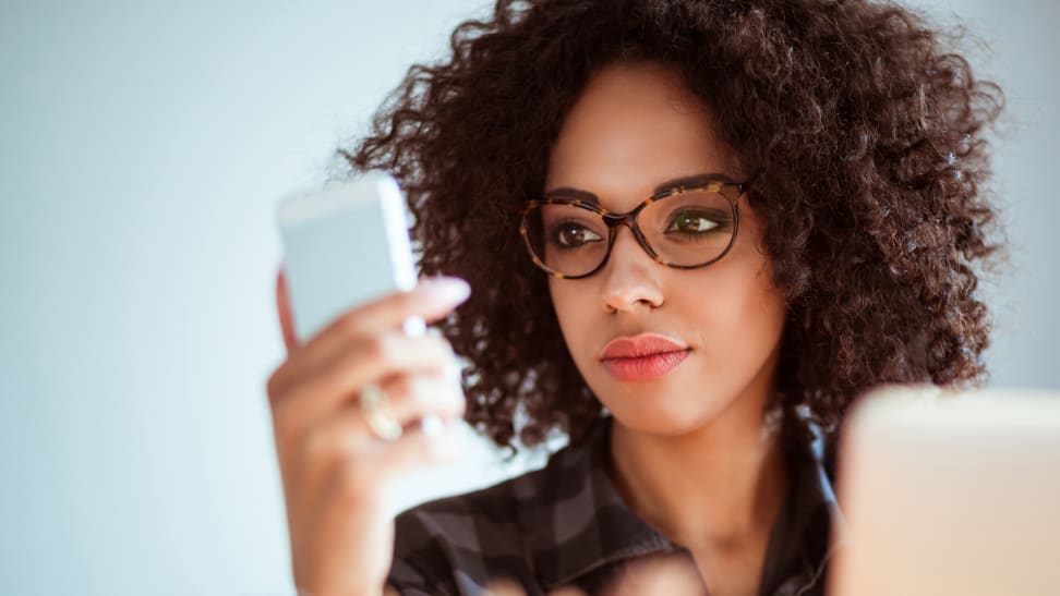 Woman with curly afro is using reading glasses to read something off of her phone.