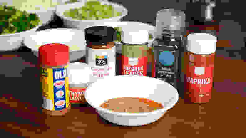 Old bay, cayenne pepper, paprika, and thyme bottles surrounded by a bowl with those ingredients mixed together.