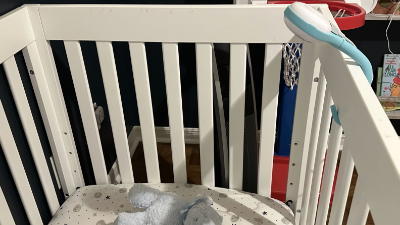 A Lollipop baby monitor is wrapped around the bars of a white crib with a mattress and stuffed animal inside.