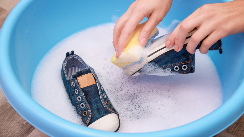 Person washing children's fabric sneakers in a plastic bin with a sponge