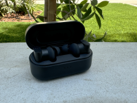 The Status Audio Between 3ANC wireless earbuds in their onyx charging case, open on a white cement wall with trees and grass in the background.