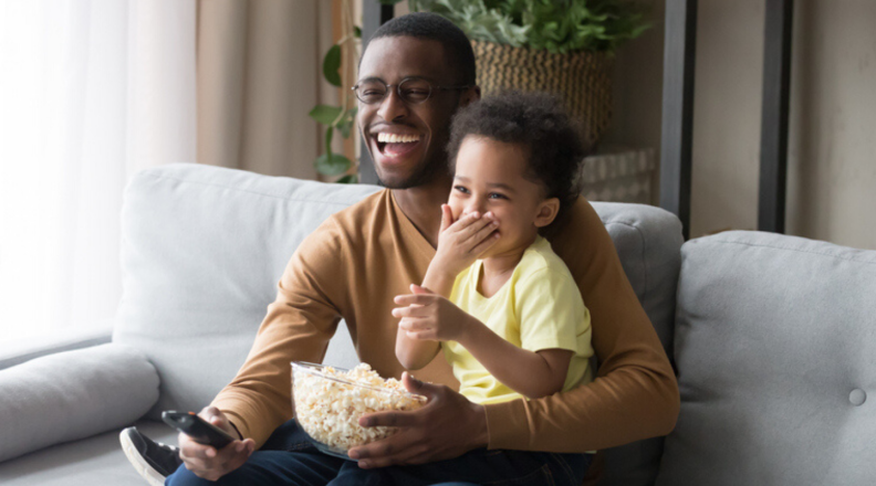 A toddler and parent sit on the couch and watch TV